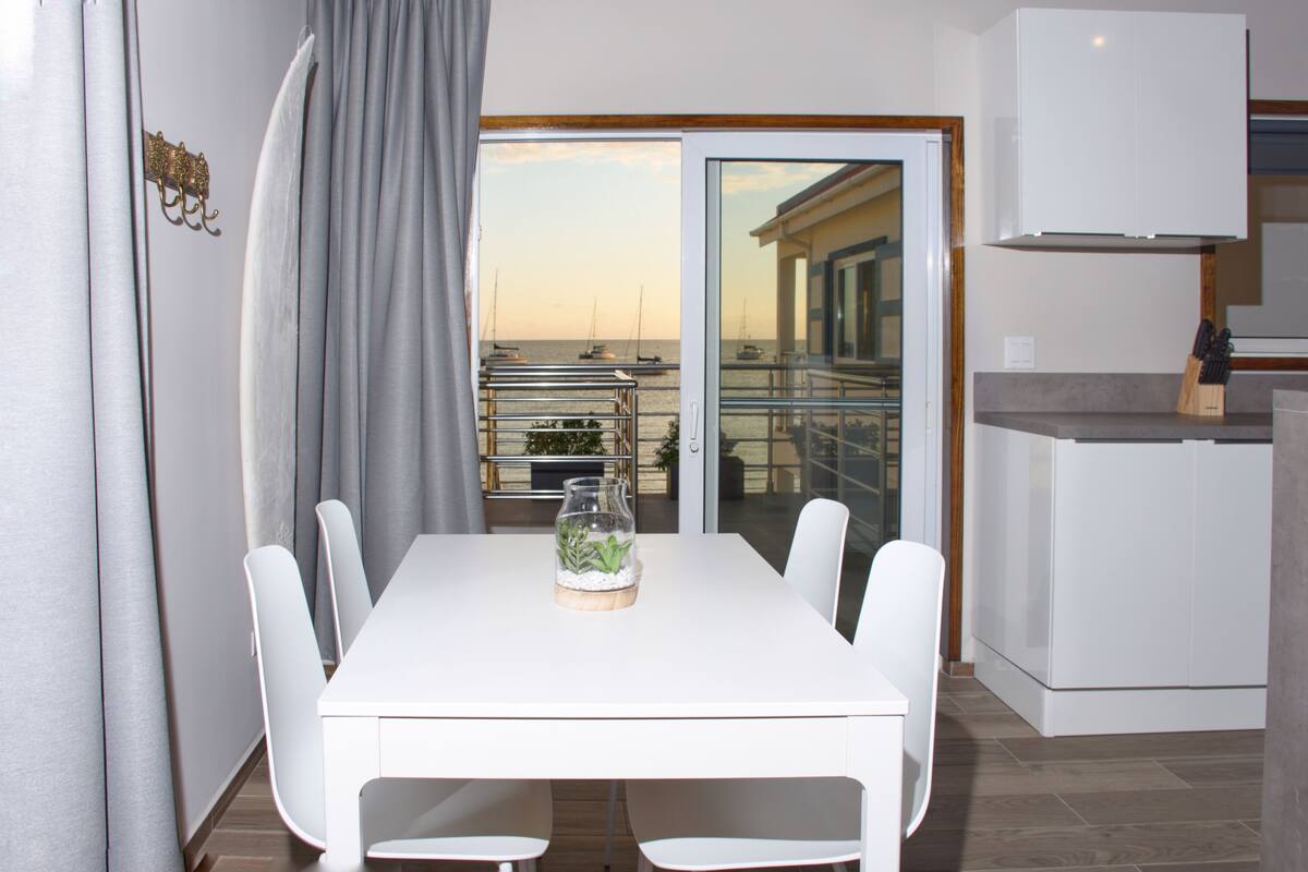A modern kitchen with a white countertop, a built-in oven, and a view of the sea through a glass door.