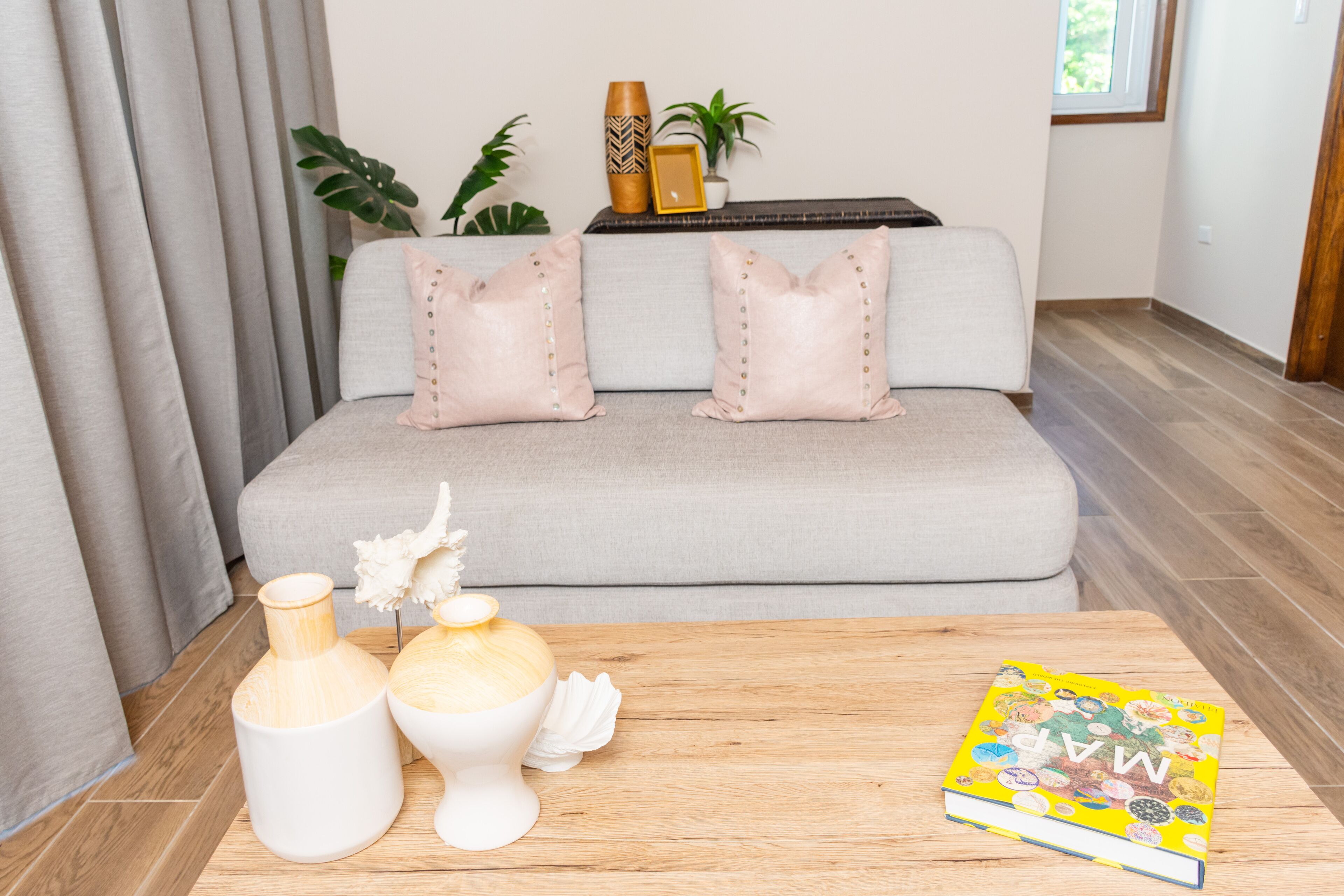 A living room with a grey sofa, two decorative pillows, a wooden coffee table with vases and a book, and a plant in the background.