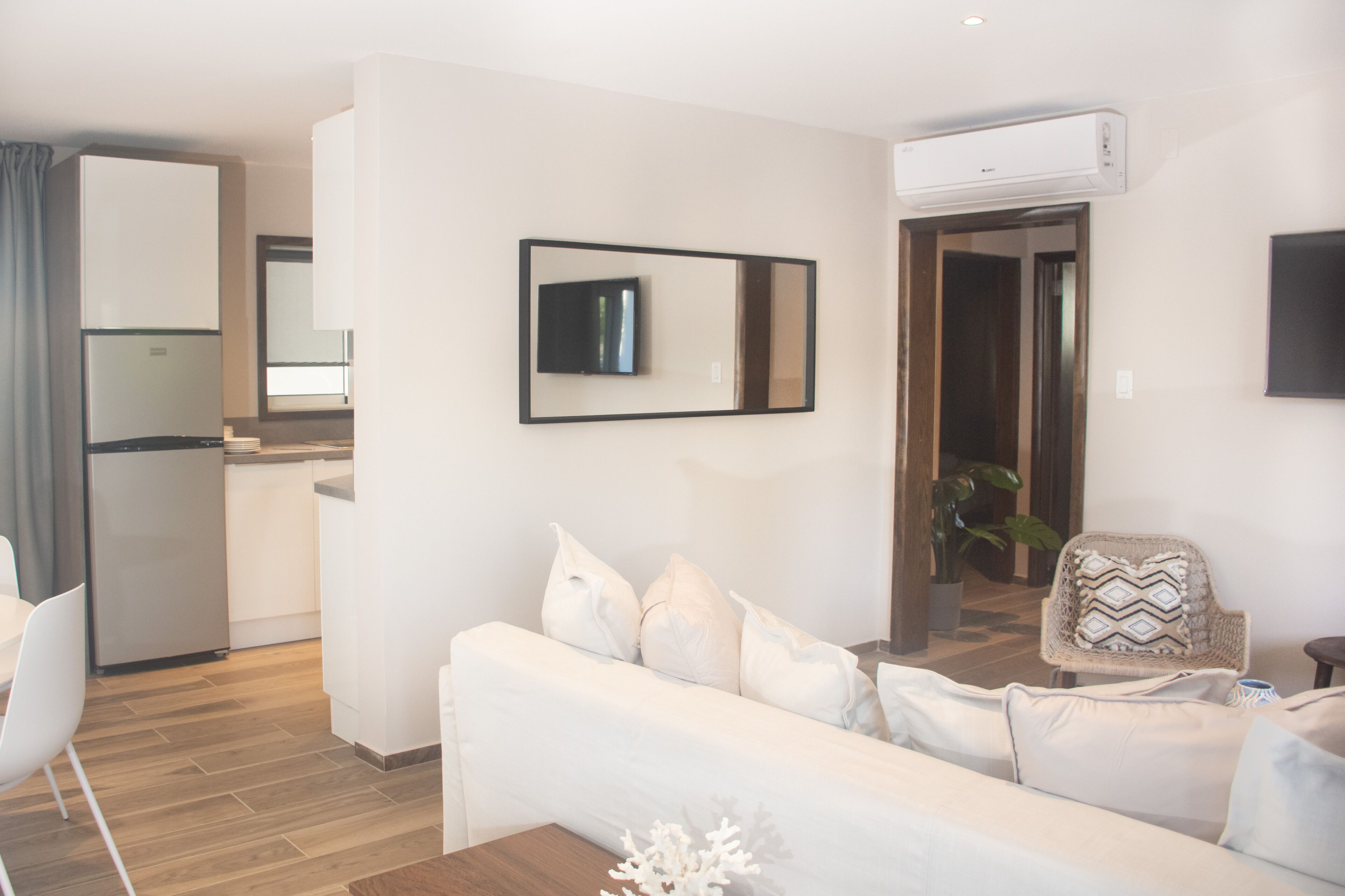 Ein modernes Wohnzimmer mit einem weißen Sofa, einem Essbereich mit Tisch und Stühlen in Weiß, einer Küche mit Kühlschrank und einem an der Wand befestigten Fernseher.