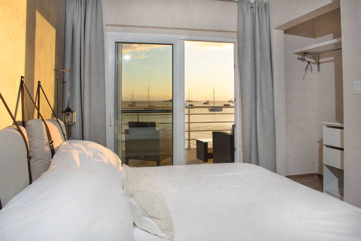 A hotel room with a bed, a chair, a table, and a view of boats on the water through an open door.