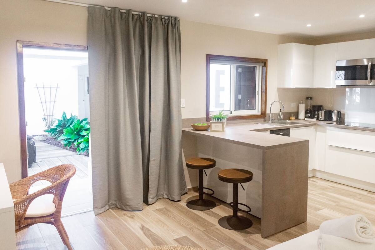 A modern kitchen with a central island, bar stools, a dining area with a wicker chair, and a view of the garden through the sliding doors.