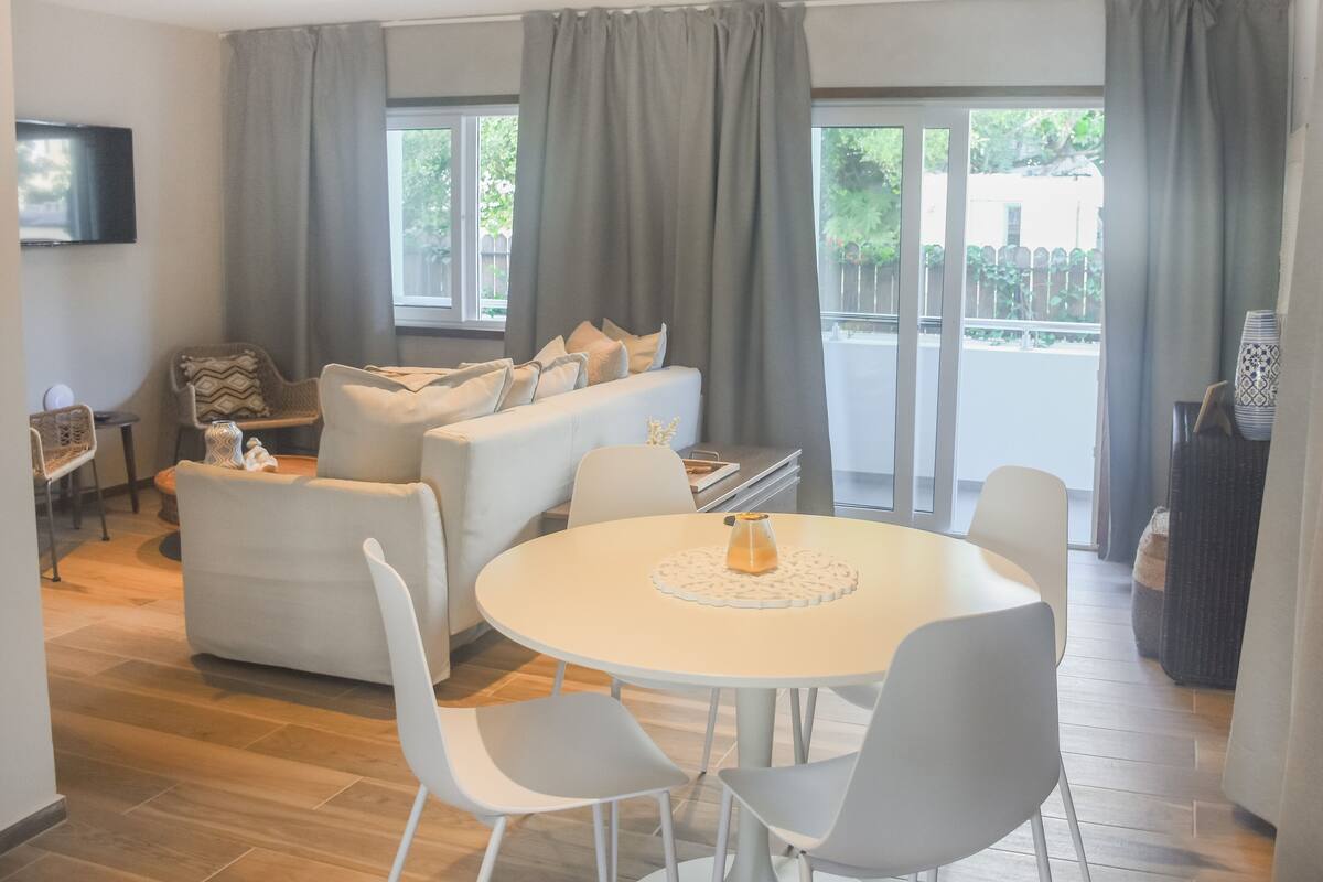A modern living room with a sofa, dining table, and chairs. There is a TV mounted on the wall and a window with curtains.