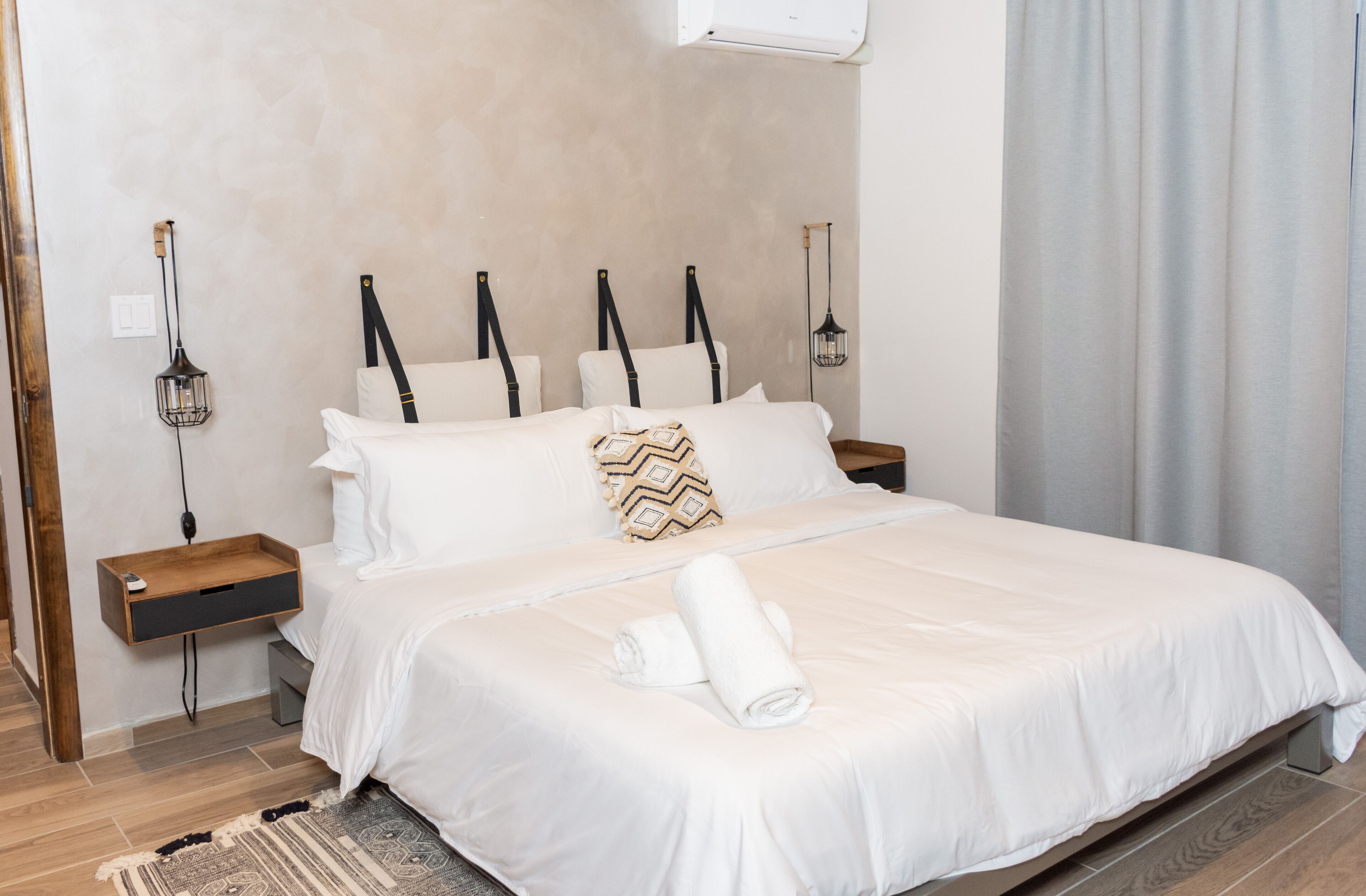 A neatly made bed with white linens, a patterned throw pillow, and rolled towels on the bed. There are two bedside tables with lamps and a power outlet. The room has a wooden floor and a light-colored wall.