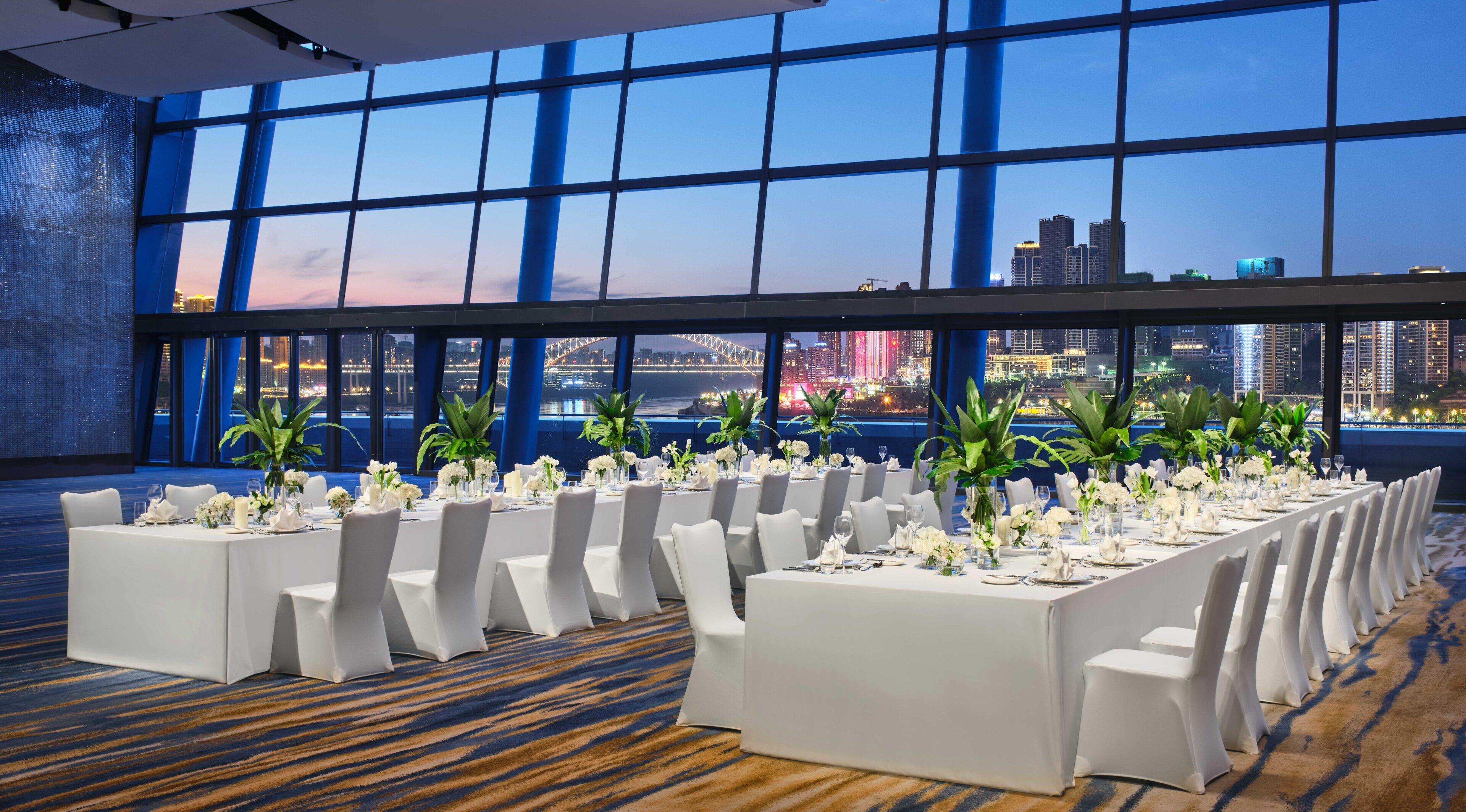 A banquet hall with tables set for a formal event, large windows offering a city skyline view, and a carpeted floor.