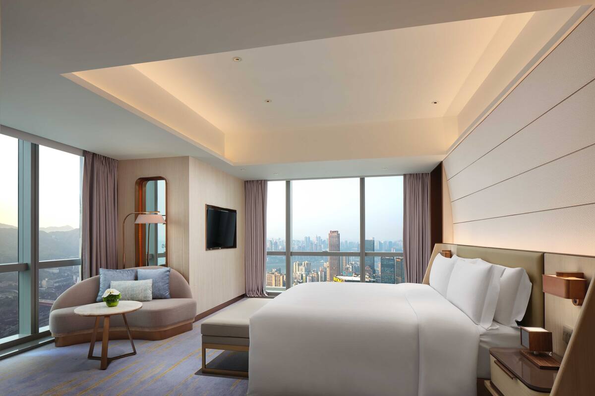 A modern hotel room with a large bed, a sofa, a small table, and a view of the cityscape.