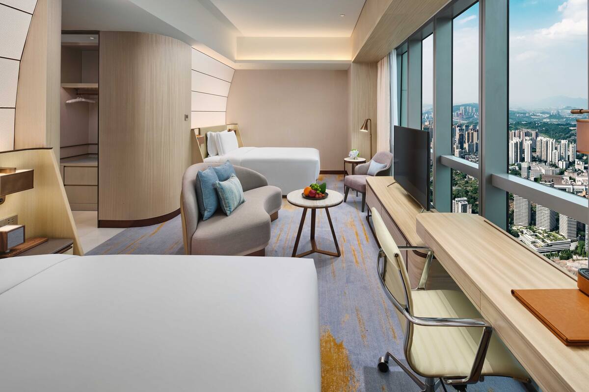 A modern hotel room with a large bed, a sofa, a desk, and a TV. The room has a city view.