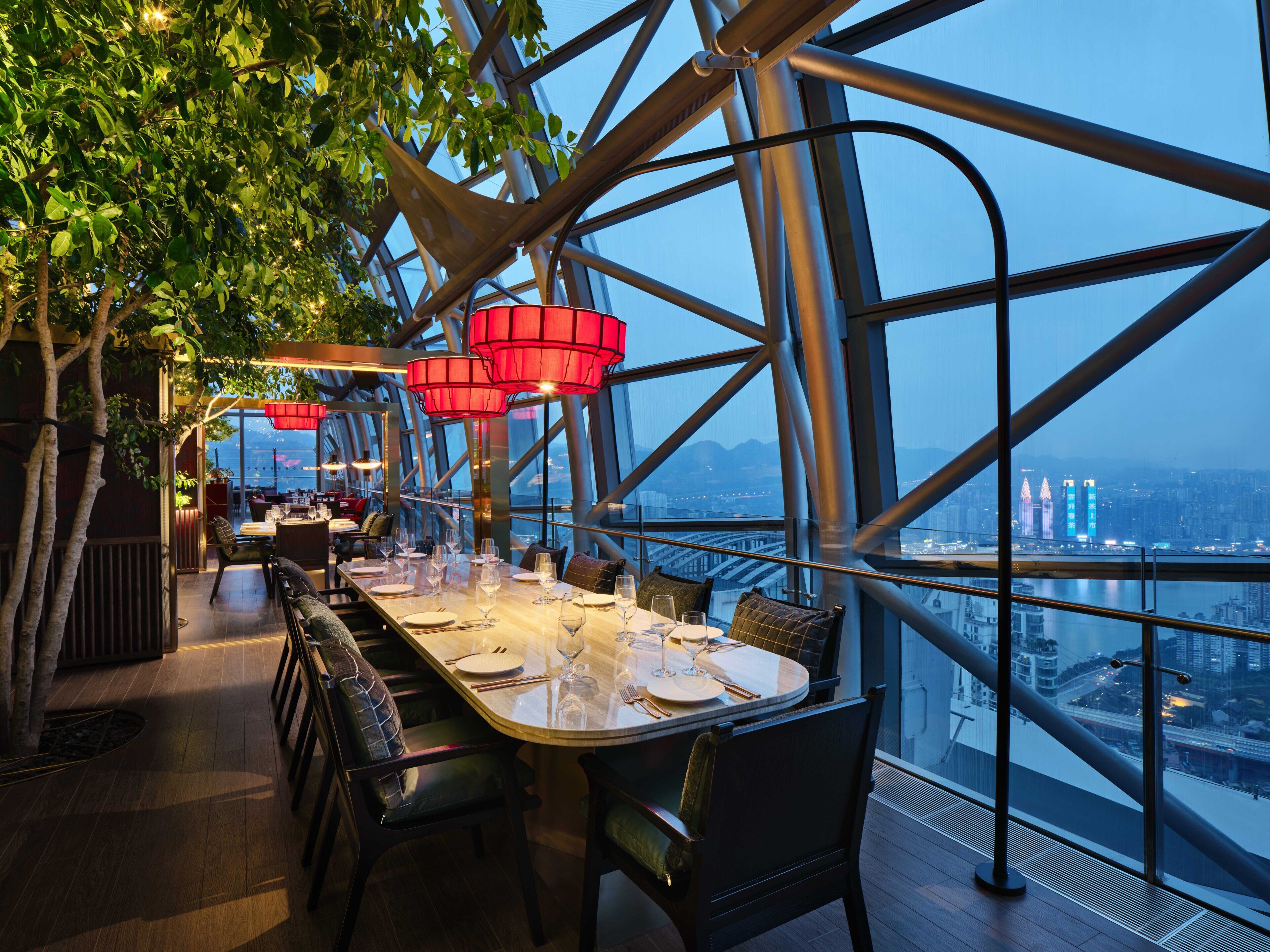 An indoor dining area with a panoramic city view, modern metal framework, and red hanging lights.