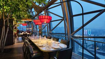 An indoor dining area with a panoramic city view, modern metal framework, and red hanging lights.
