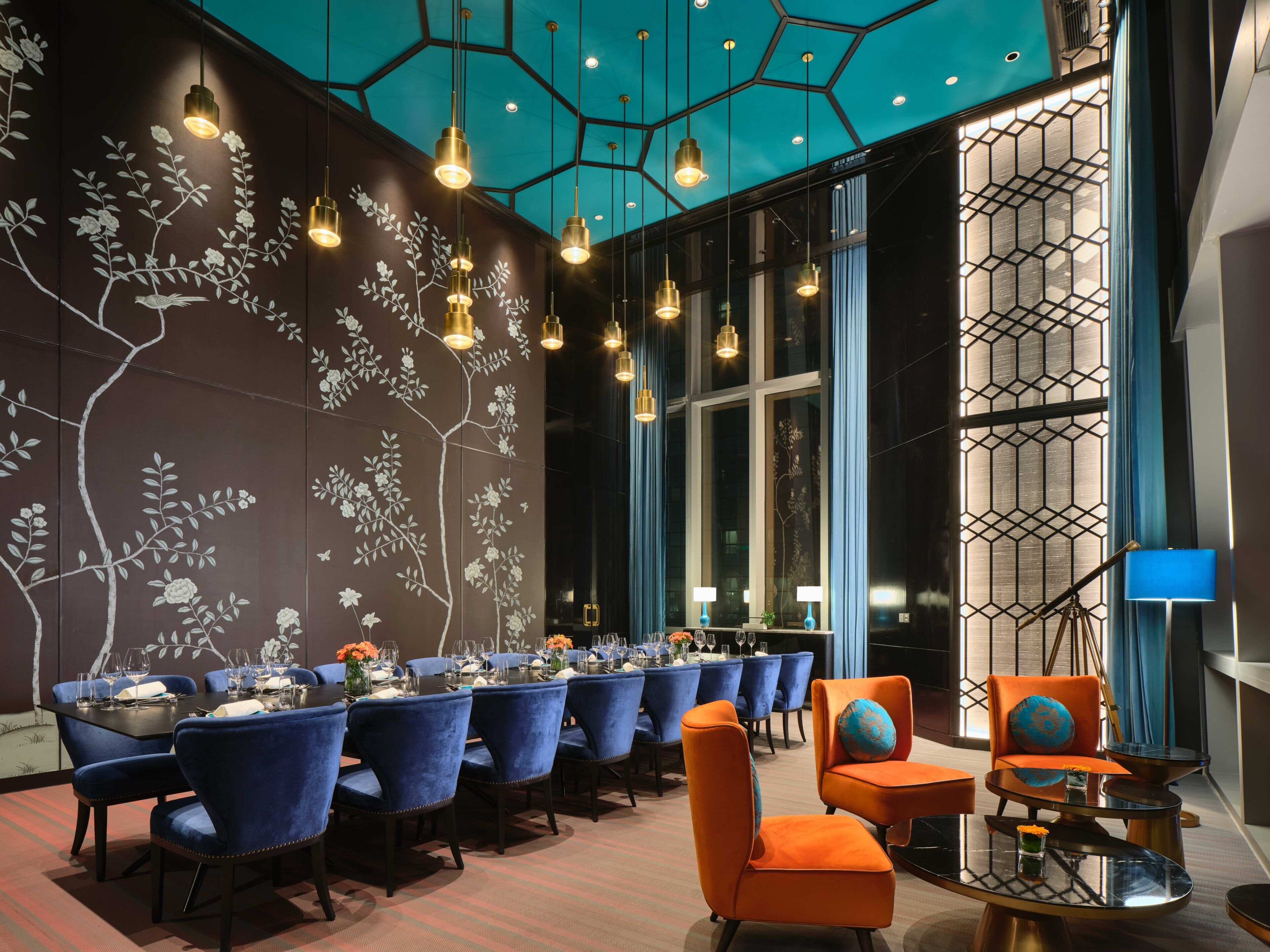 Un espace repas avec une longue table dressée pour un repas, des chaises à l’assise rembourrée de couleur bleue et un design moderne au niveau du plafond.