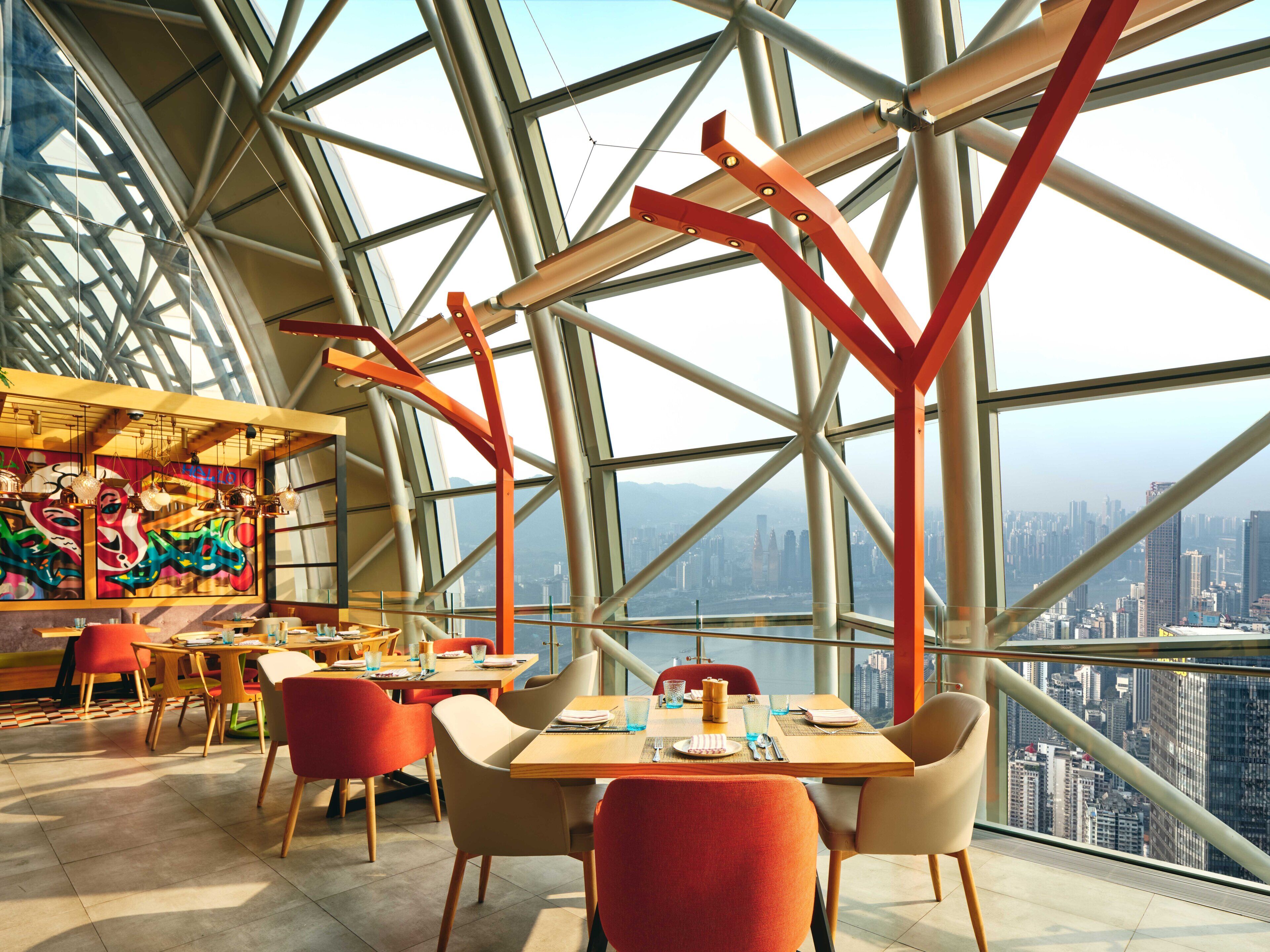 A high-rise restaurant with a panoramic city view, featuring a modern interior with geometric ceiling design and colorful artwork.