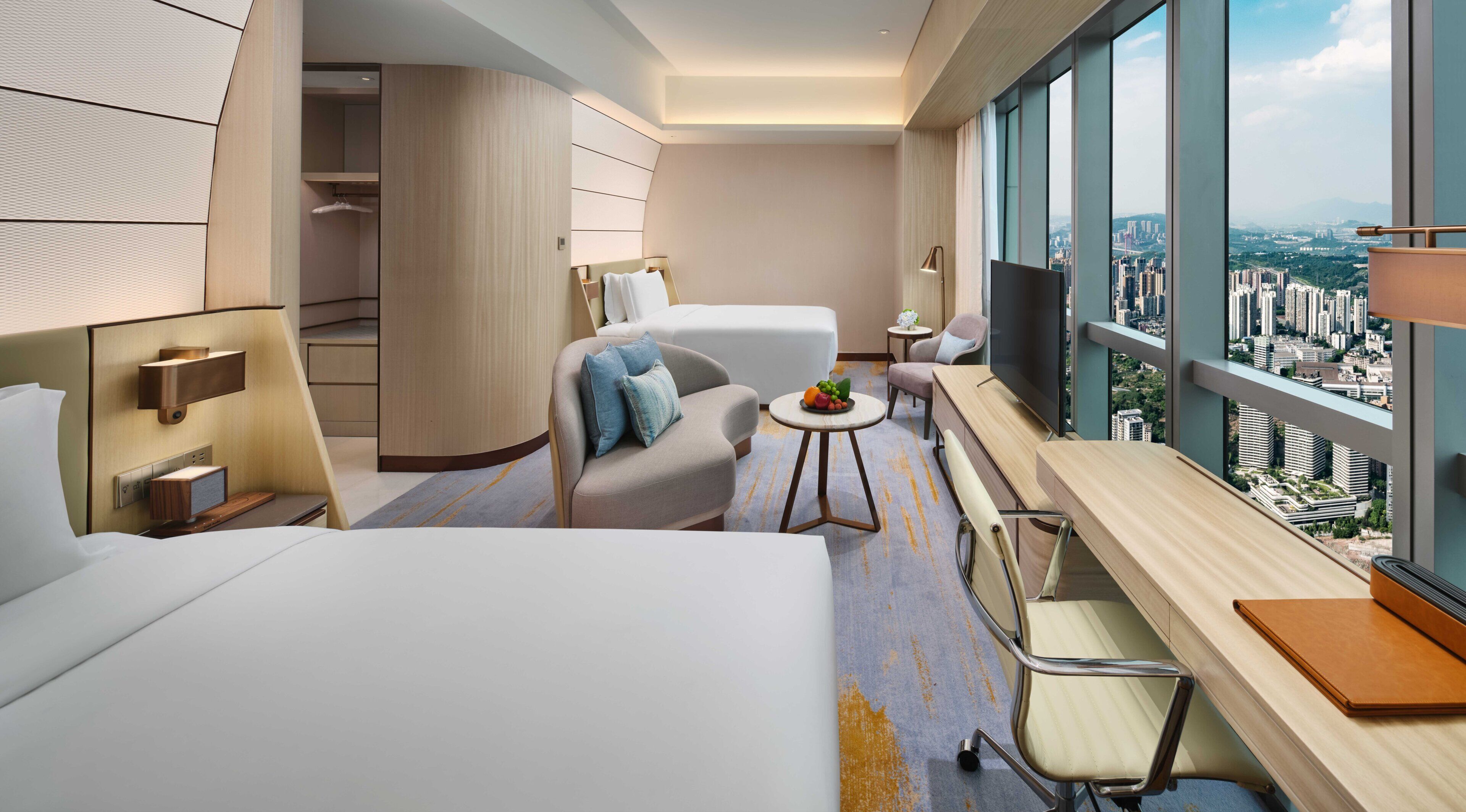 A hotel room with a bed, a sofa, a desk, a TV, and a view of the city.
