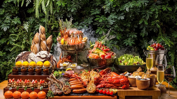 Una mesa con diversos alimentos, entre ellos mariscos, pan y frutas, frente a un exuberante follaje.
