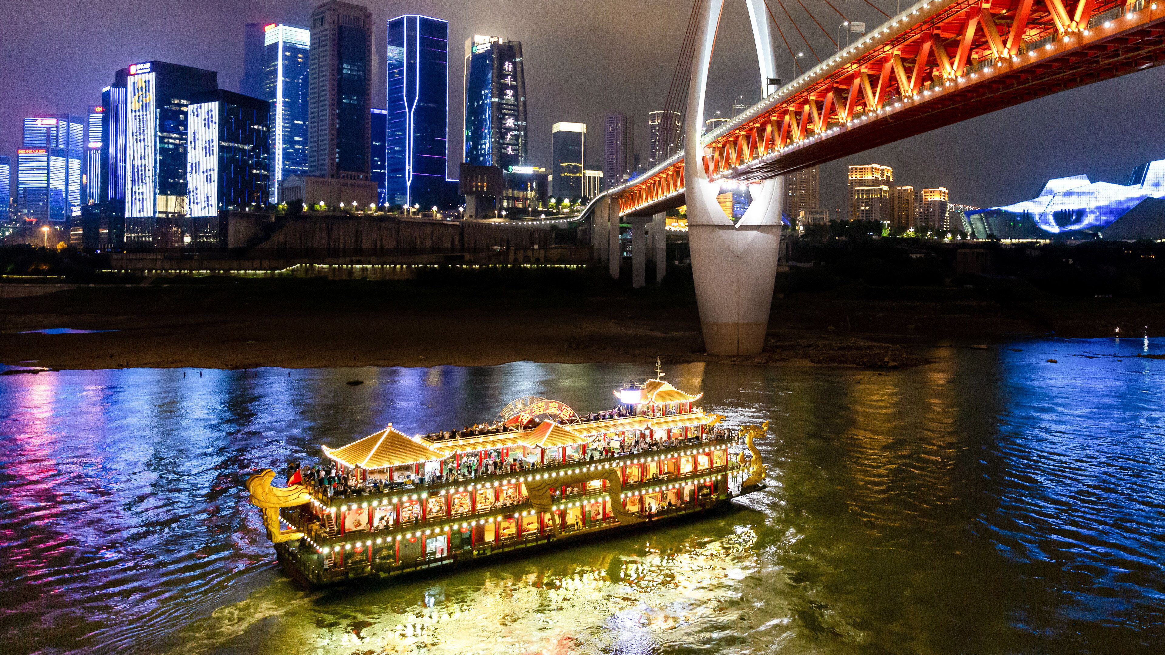 A riverboat with multiple decks and a large, illuminated structure in the background.