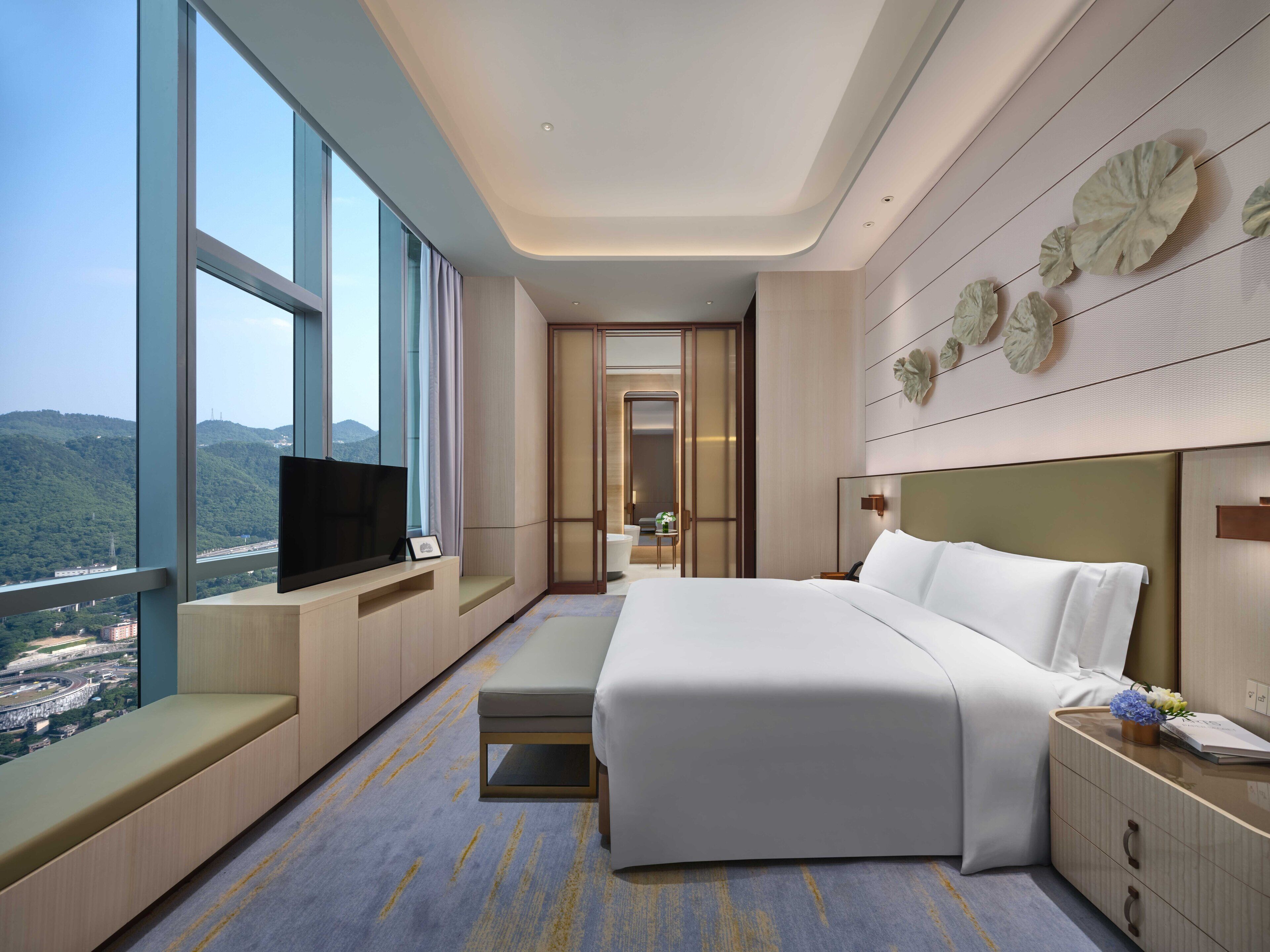 A modern hotel room with a large bed, a TV on a built-in bench, a view of the city, and a decorative wall panel.