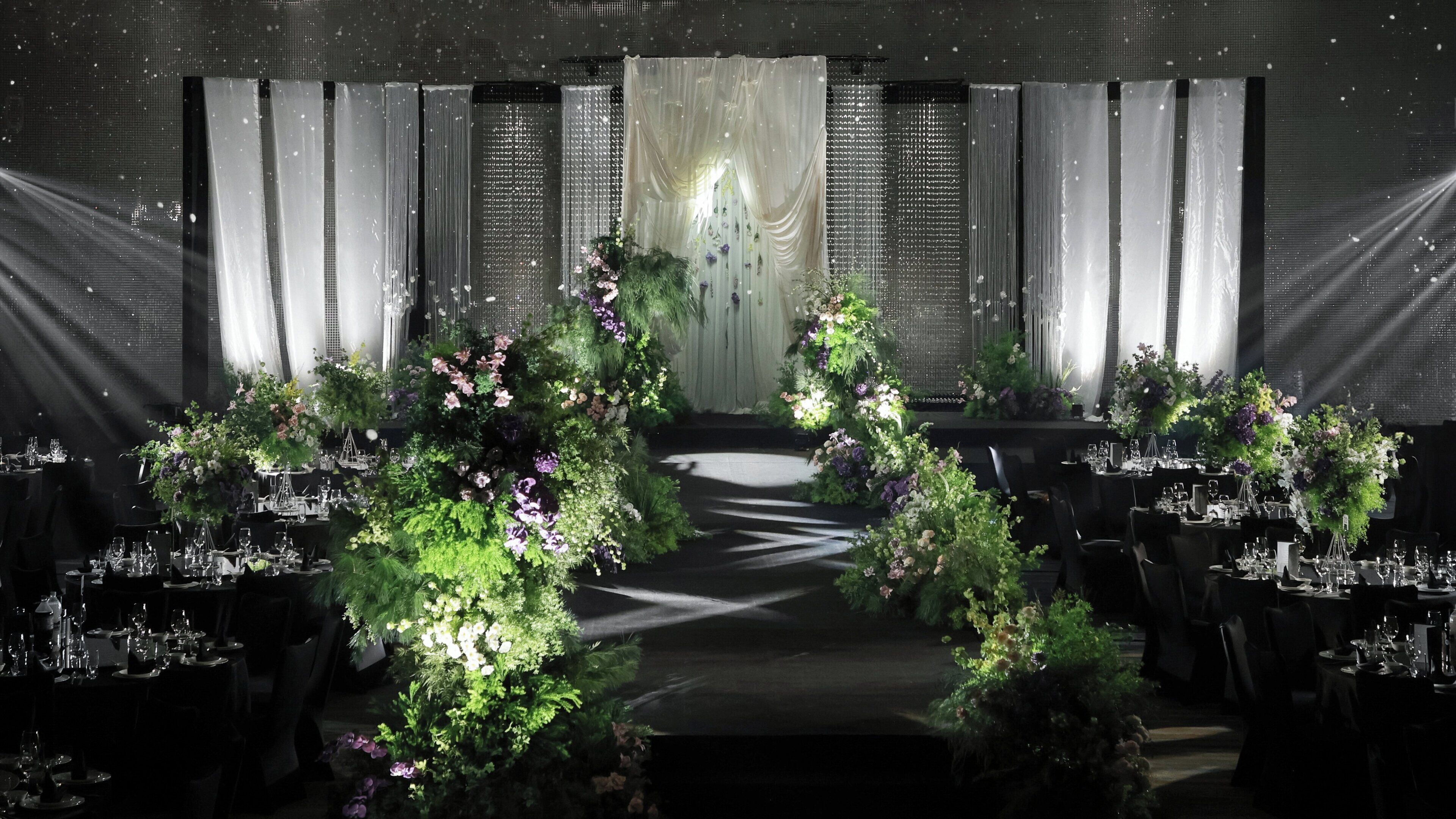 A formal dining area with tables set for a meal, a stage with curtains, and floral arrangements.