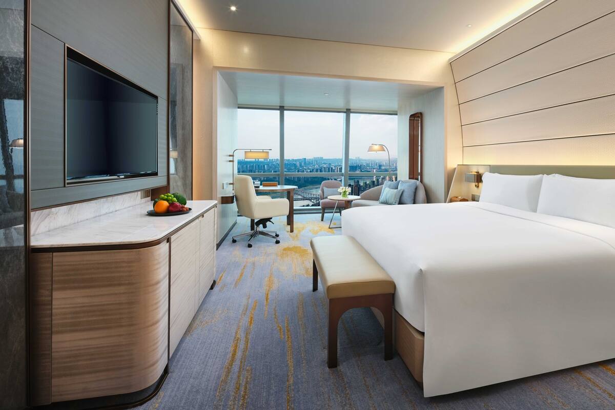 A modern hotel room with a large bed, a desk with a flat-screen TV, a seating area with a view of the city, and a large window.
