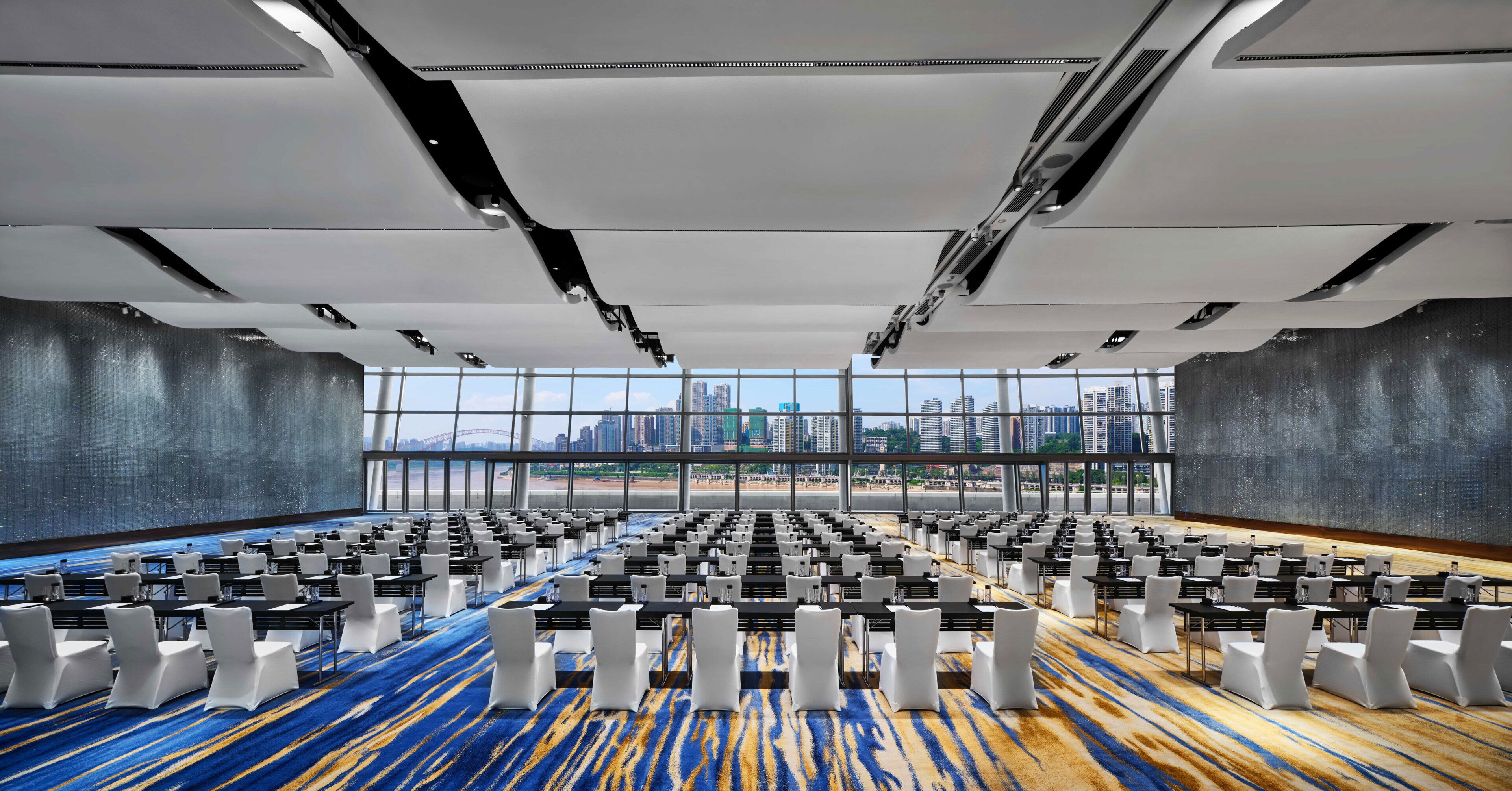 Une salle de conférence avec des rangées de tables et de chaises, de grandes fenêtres donnant sur la ville et un tapis à motifs.
