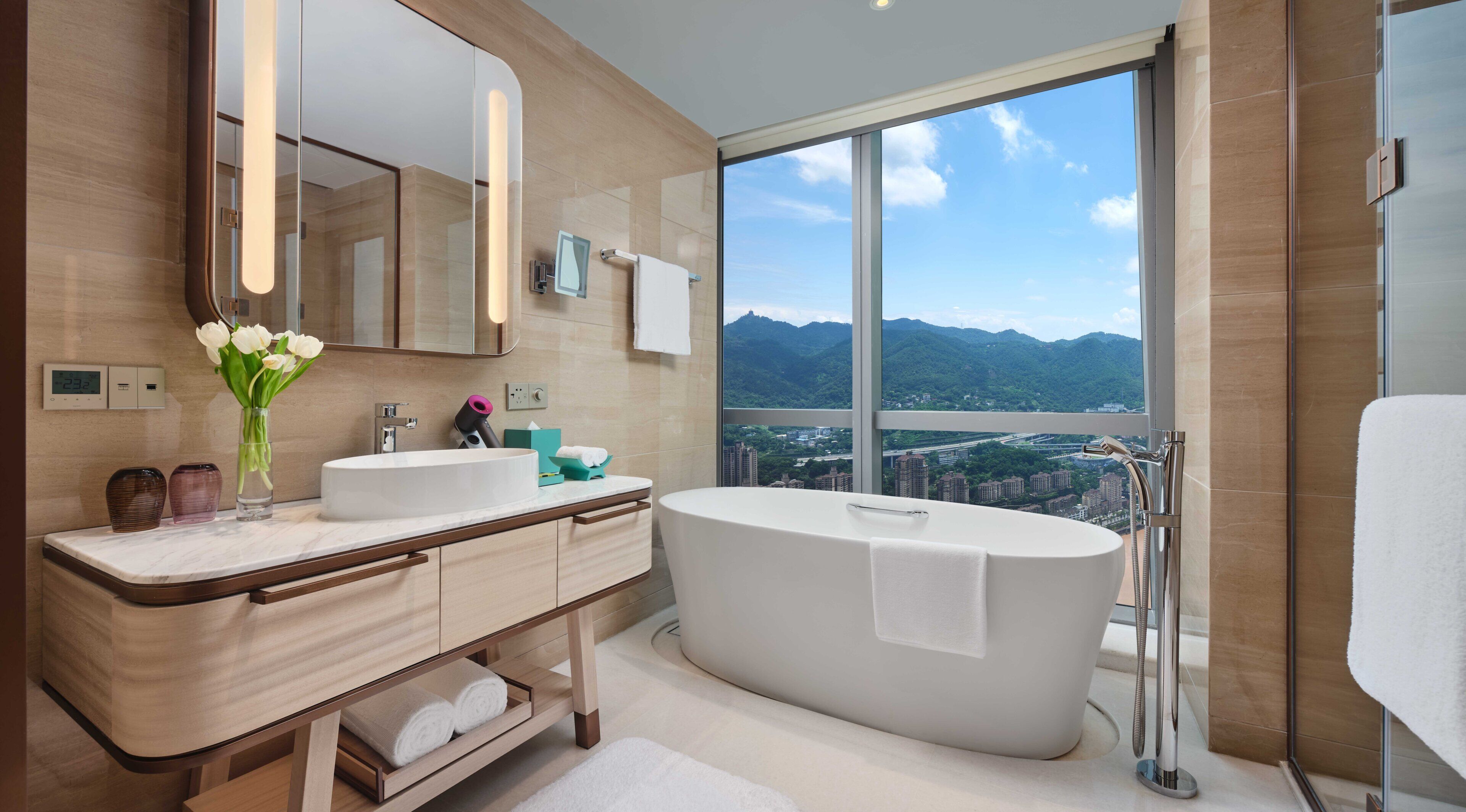 Une salle de bain moderne dotée d’une grande baignoire, d’un meuble-lavabo à deux vasques, d’un miroir et offrant une vue sur le paysage urbain et les montagnes grâce à de grandes fenêtres.