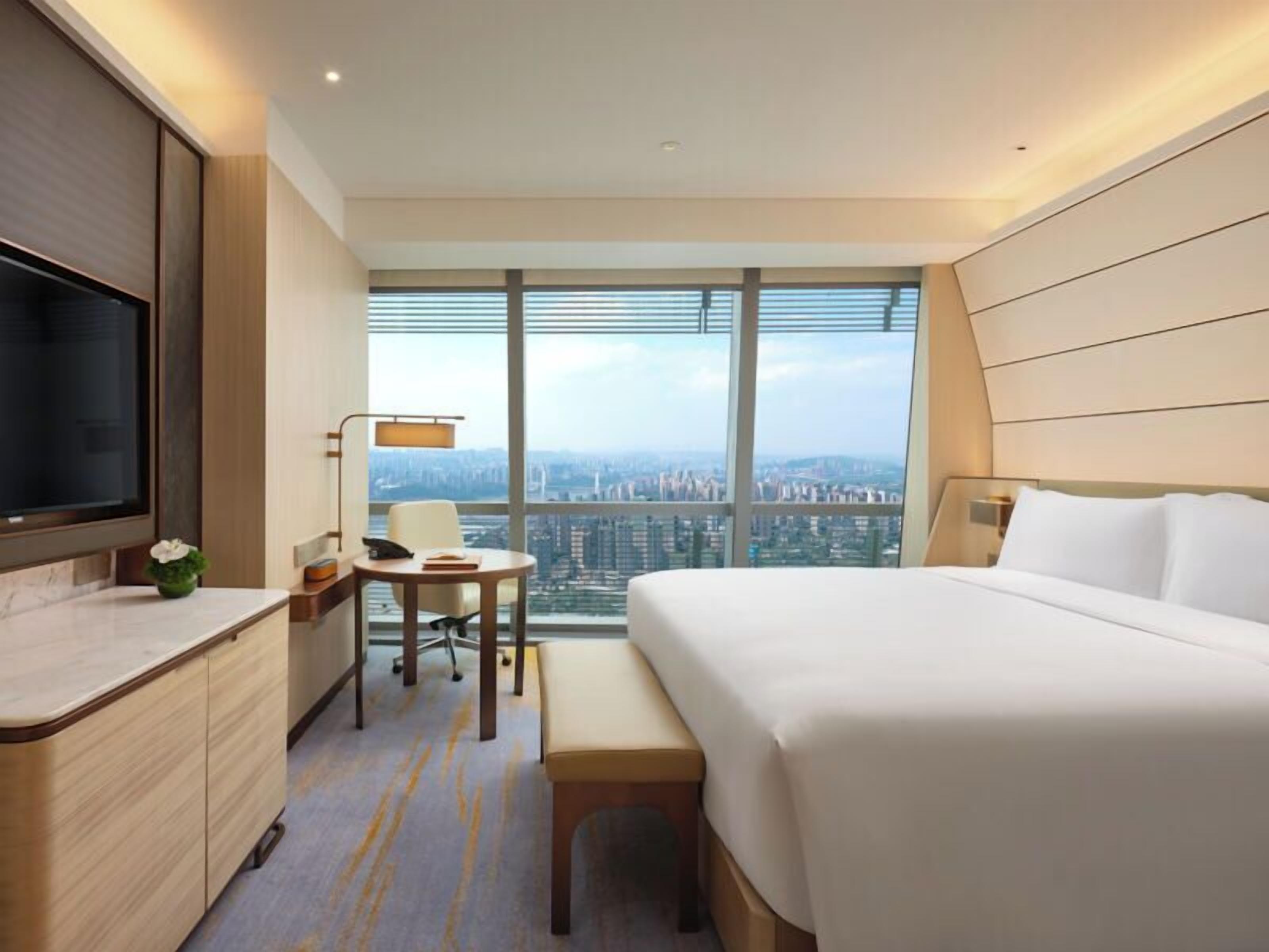 A hotel room with a bed, desk, chair, and a large window offering a city view.