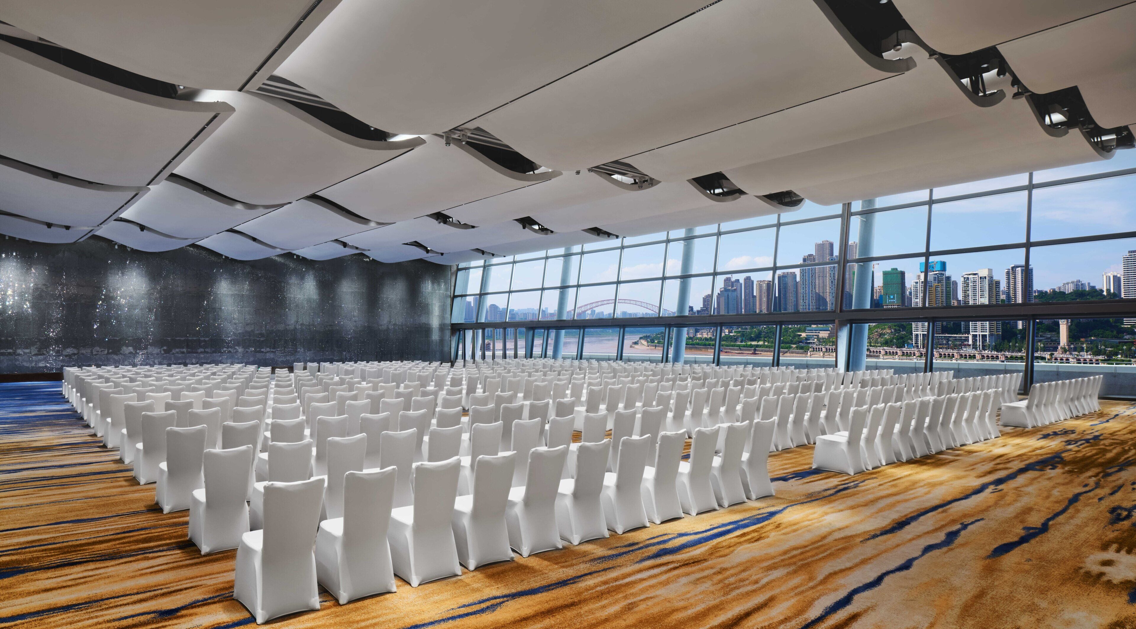 A large conference room with rows of white chairs, a carpeted floor, and a cityscape view through large windows.