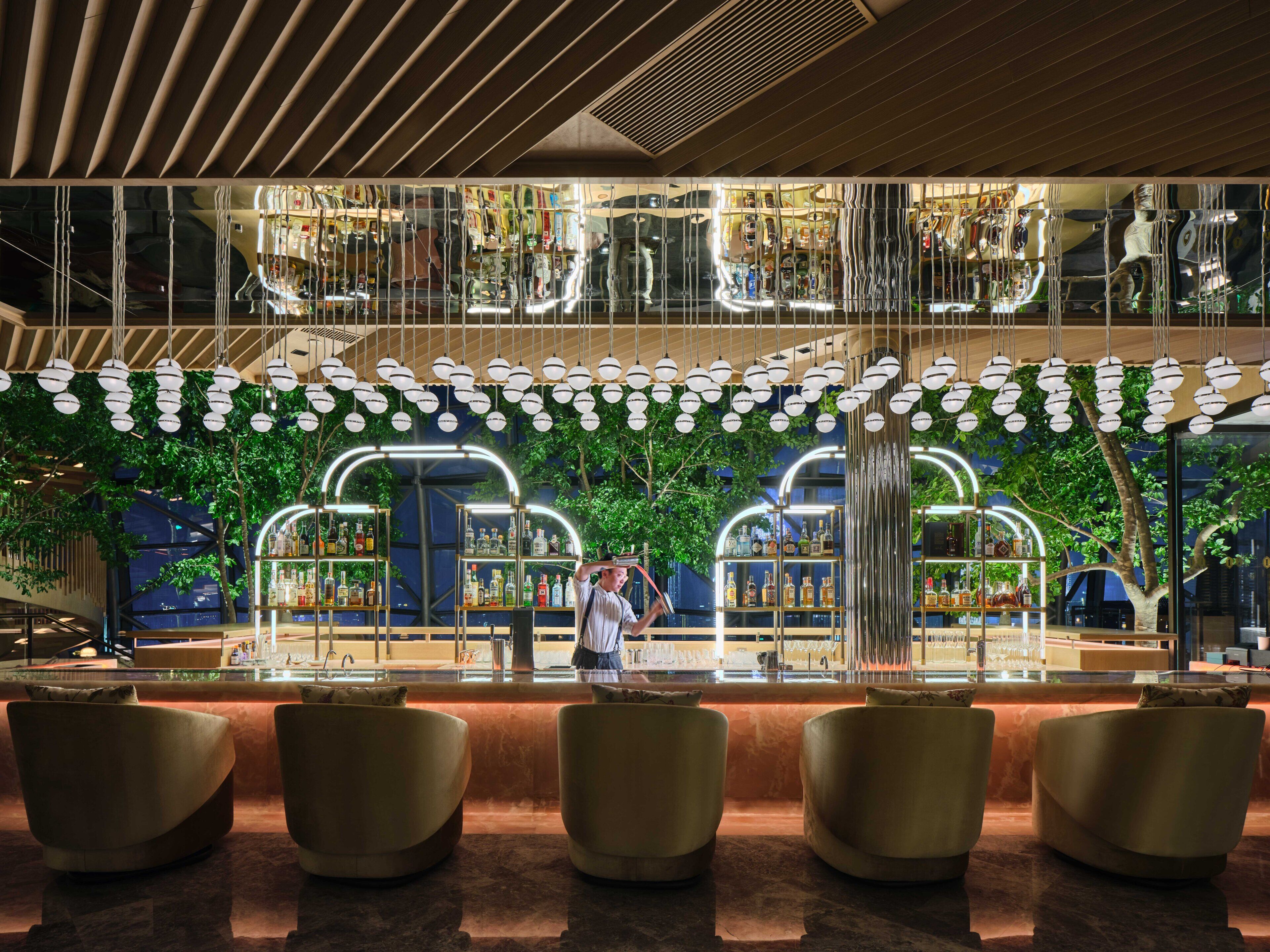 Un área de bar moderna con una partición de vidrio, luces esféricas colgantes y un espacio de asiento con sillones acolchados.