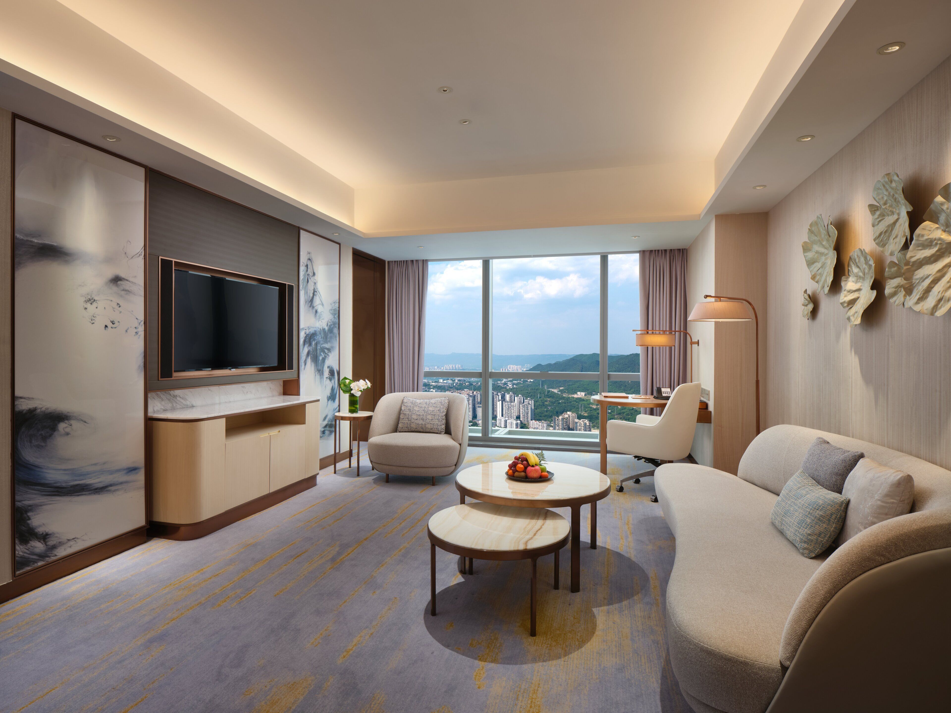 A modern hotel room with a large TV, comfortable seating, a dining area, and a view of the city.