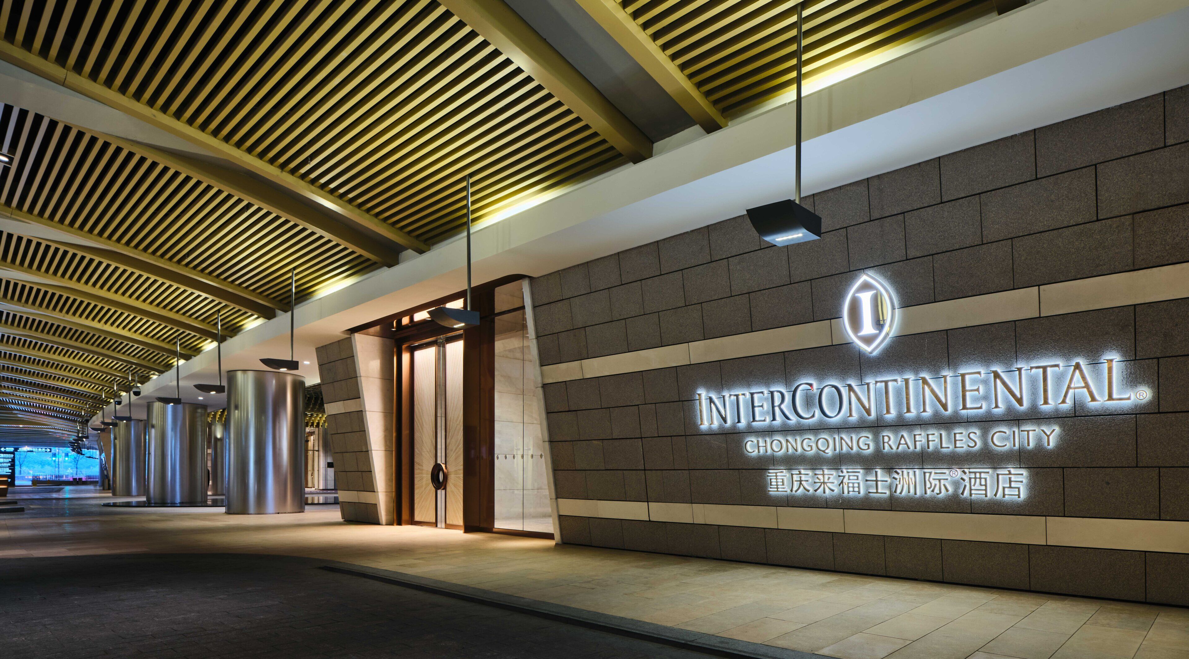 A corridor with a modern ceiling design, a wall with illuminated signage for Intercontinental Chongqing Raffles City, and a door leading to a room.