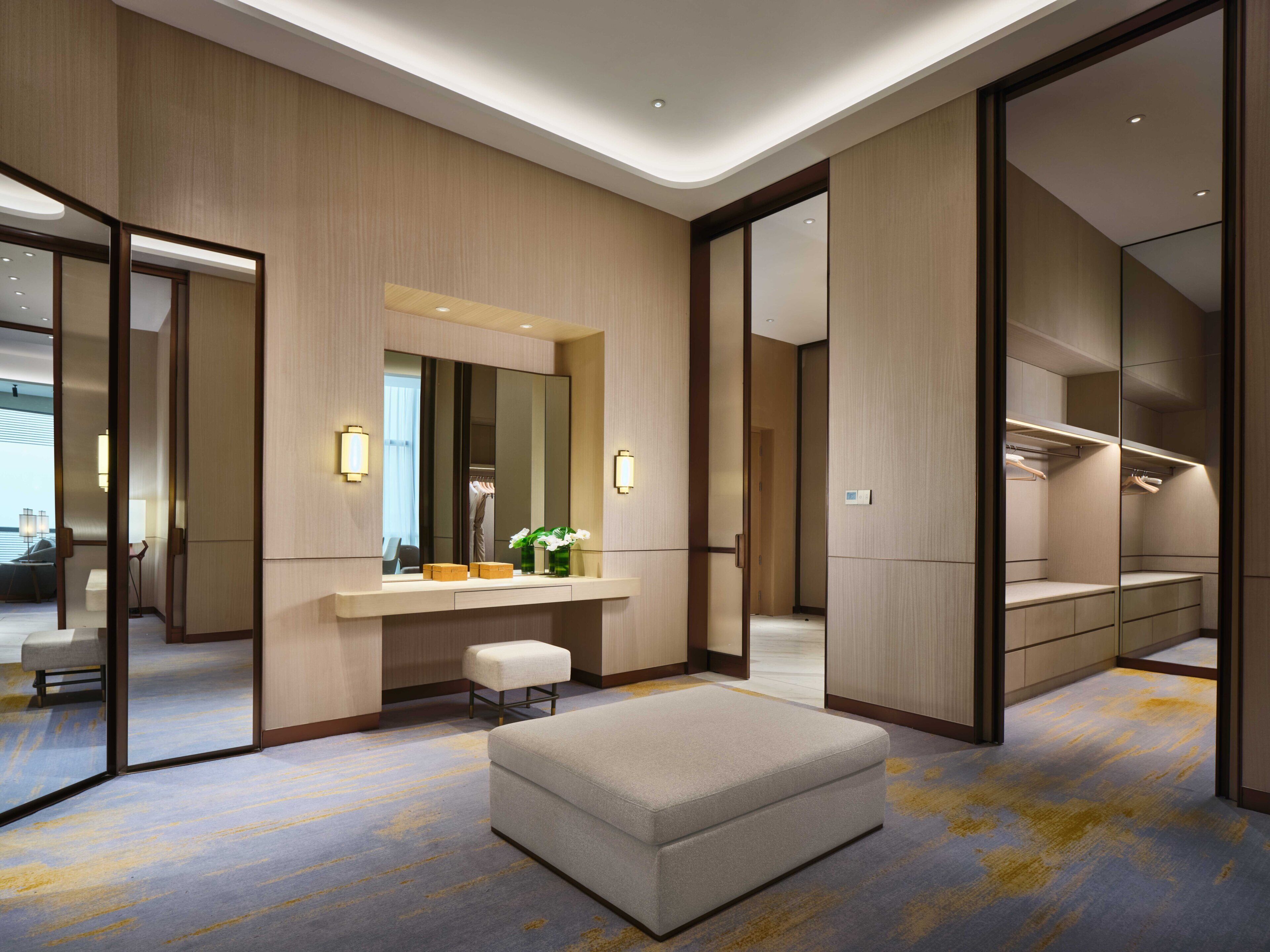 Une chambre d’hôtel moderne, dotée d’un grand miroir, d’une coiffeuse avec miroir, d’un petit tabouret et d’un banc rembourré.