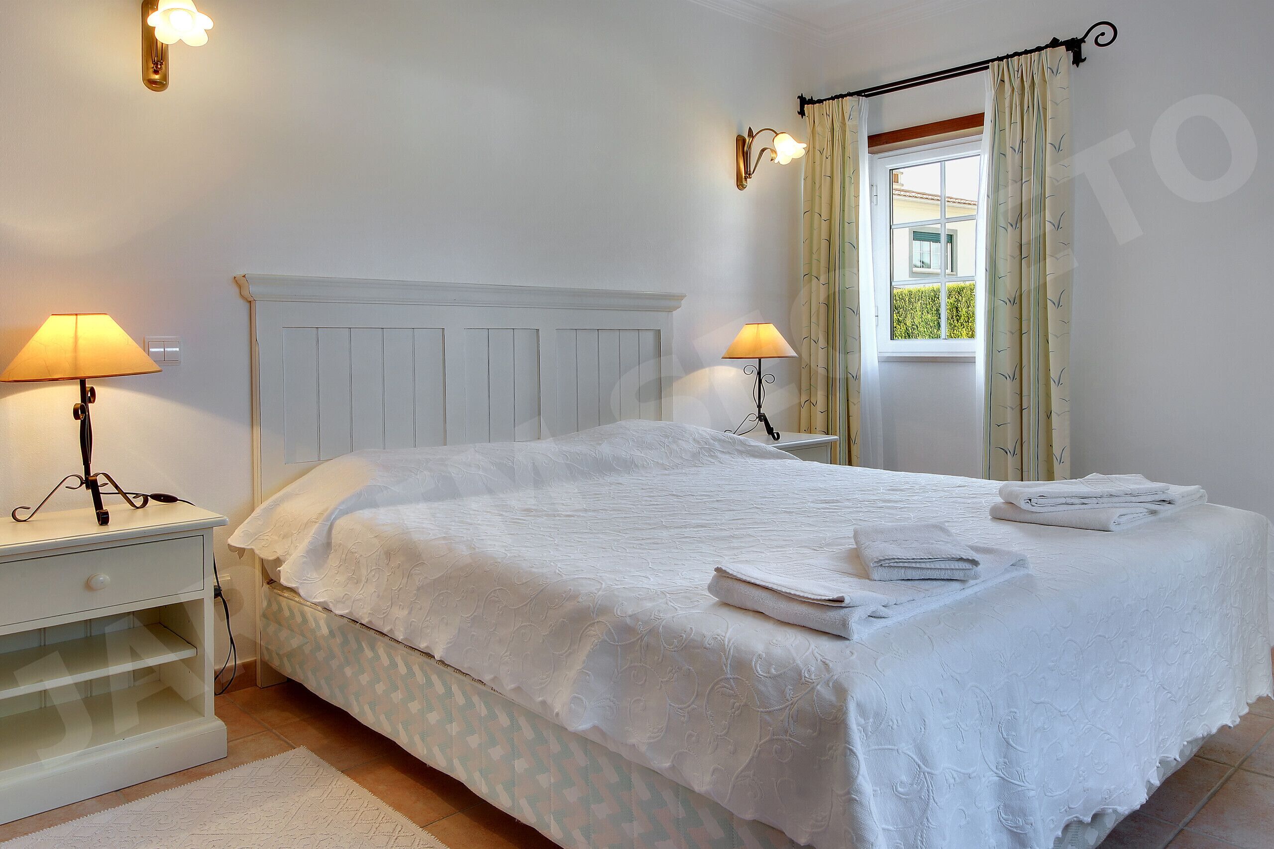 A neatly made bed with white linens, two bedside lamps, and a window with curtains.