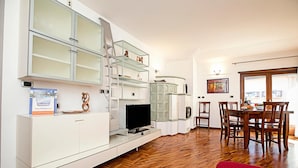 Una sala de estar moderna con televisor, muebles blancos, una zona de comedor con sillas de madera y un sofá rojo.