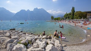 A rocky beach with clear water, mountains in the background, and people swimming and relaxing. - Bella Arco Apartment with Balcony, Arco, Italy (Arco)