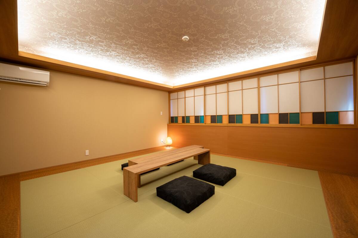 A tatami mat room with a low wooden bench and cushions, a ceiling fan, and a shoji screen with a geometric pattern.