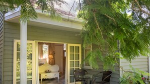 A cozy outdoor seating area with a glass table and wicker chairs, a white bench with a floral cushion, and a door leading to a room with a bed and a lamp. - Historic Foxglove Open Gardens minutes from beach and walking trails (Tilba Tilba)