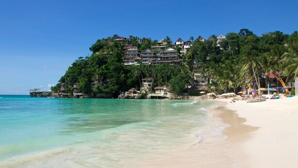 Une plage à l’eau turquoise et limpide, un rivage sablonneux et un complexe hôtelier composé de plusieurs bâtiments, le tout entouré d’une végétation luxuriante.