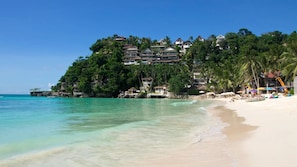 Une plage à l’eau turquoise et limpide, un rivage sablonneux et un complexe hôtelier composé de plusieurs bâtiments, le tout entouré d’une végétation luxuriante.