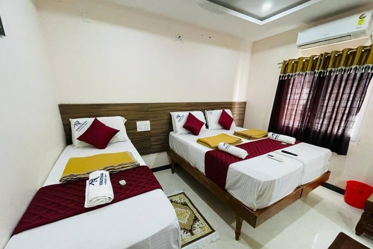 A hotel room with two single beds, each with a wooden headboard, white bedding, and red and yellow pillows. There is a red bucket and a brown stool in the corner.