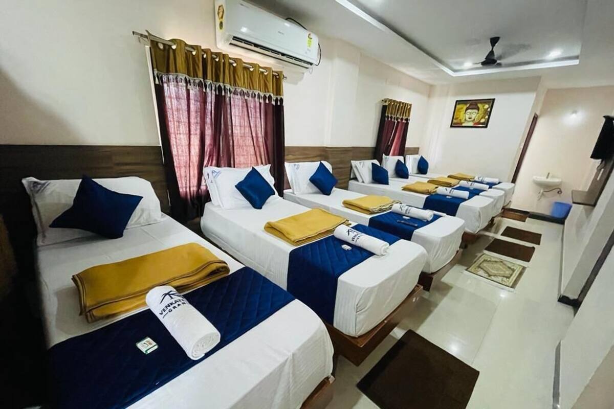 A hotel room with four single beds, each with a blue and white duvet, arranged in a row. There is a small table with a yellow towel and a white bottle, and a ceiling fan above the beds.