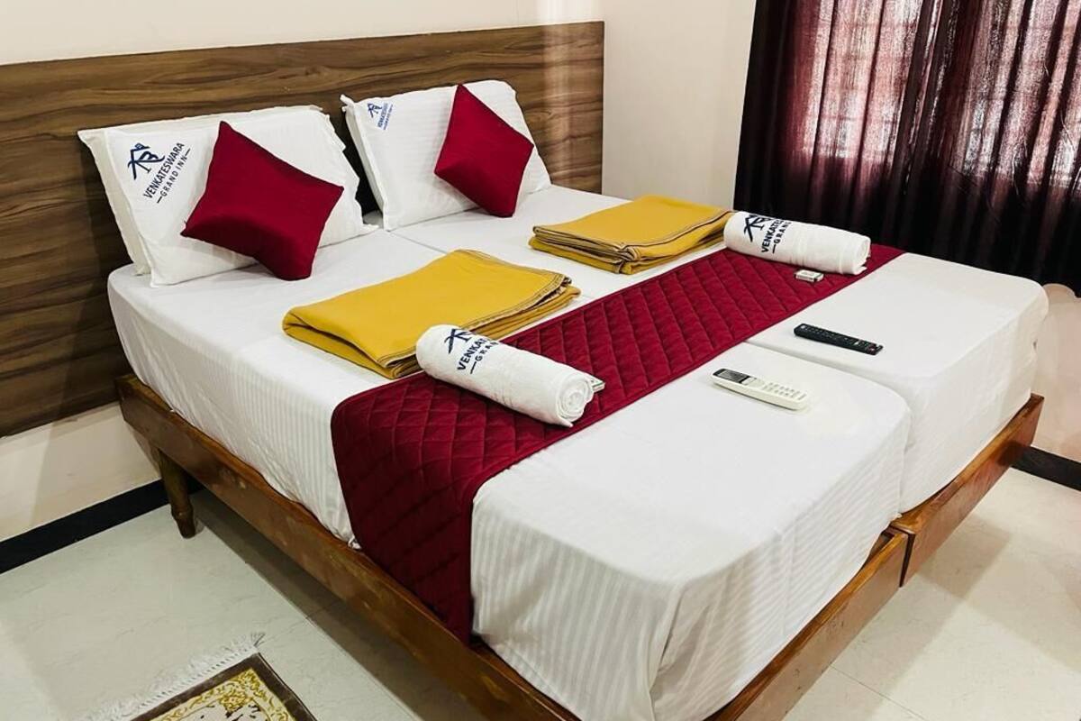 A neatly made double bed with a wooden headboard, white bedding, red and yellow accents, and a small bedside table with a remote control.