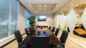 Un salón de conferencias con una larga mesa, sillas negras, una planta y un televisor en la pared.