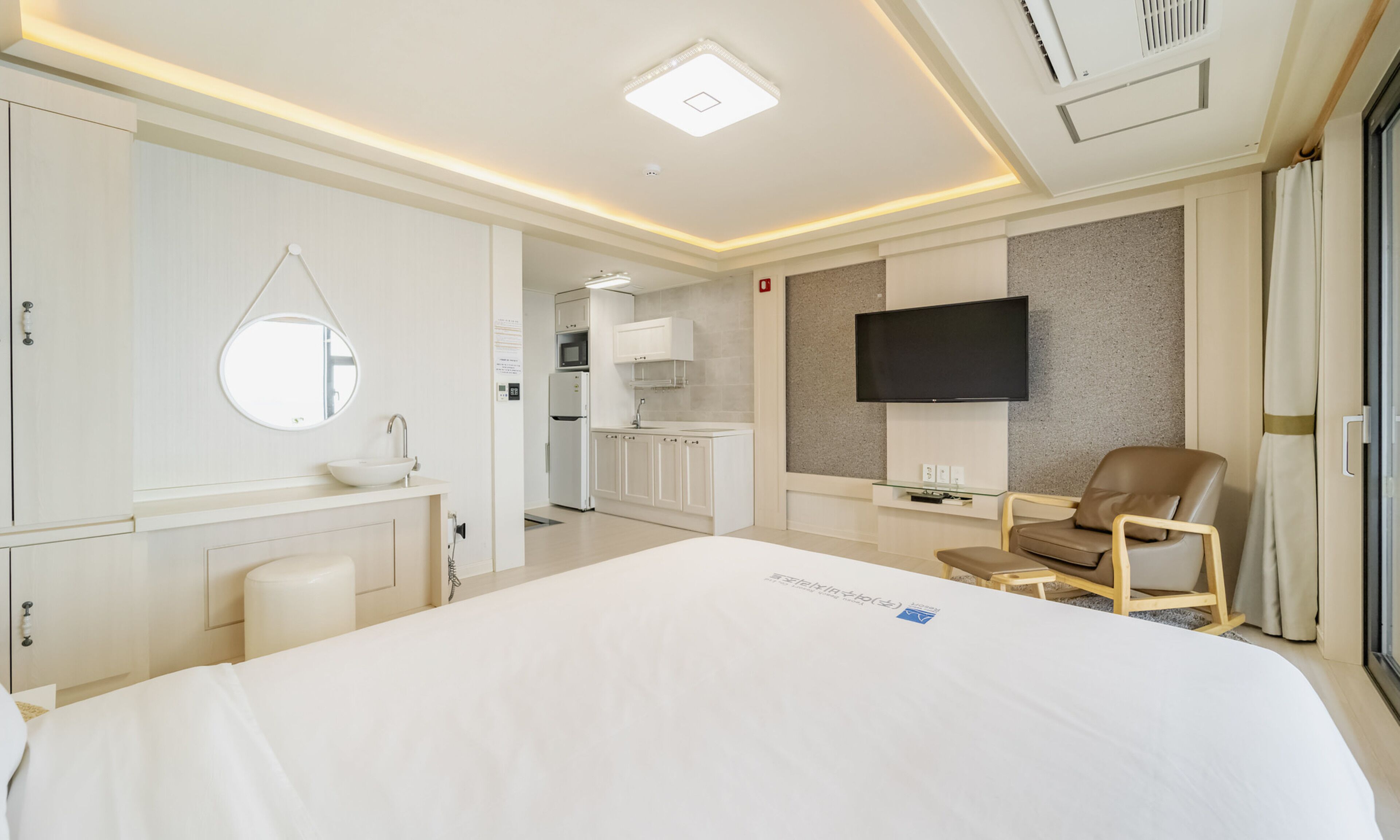 Habitación de hotel con una cama grande, un espejo redondo, un televisor de pantalla plana montado en la pared, una silla de madera y una kitchenette al fondo.