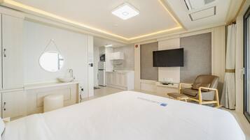 A hotel room with a large bed, a round mirror, a flat-screen TV mounted on the wall, a wooden chair, and a kitchenette in the background.