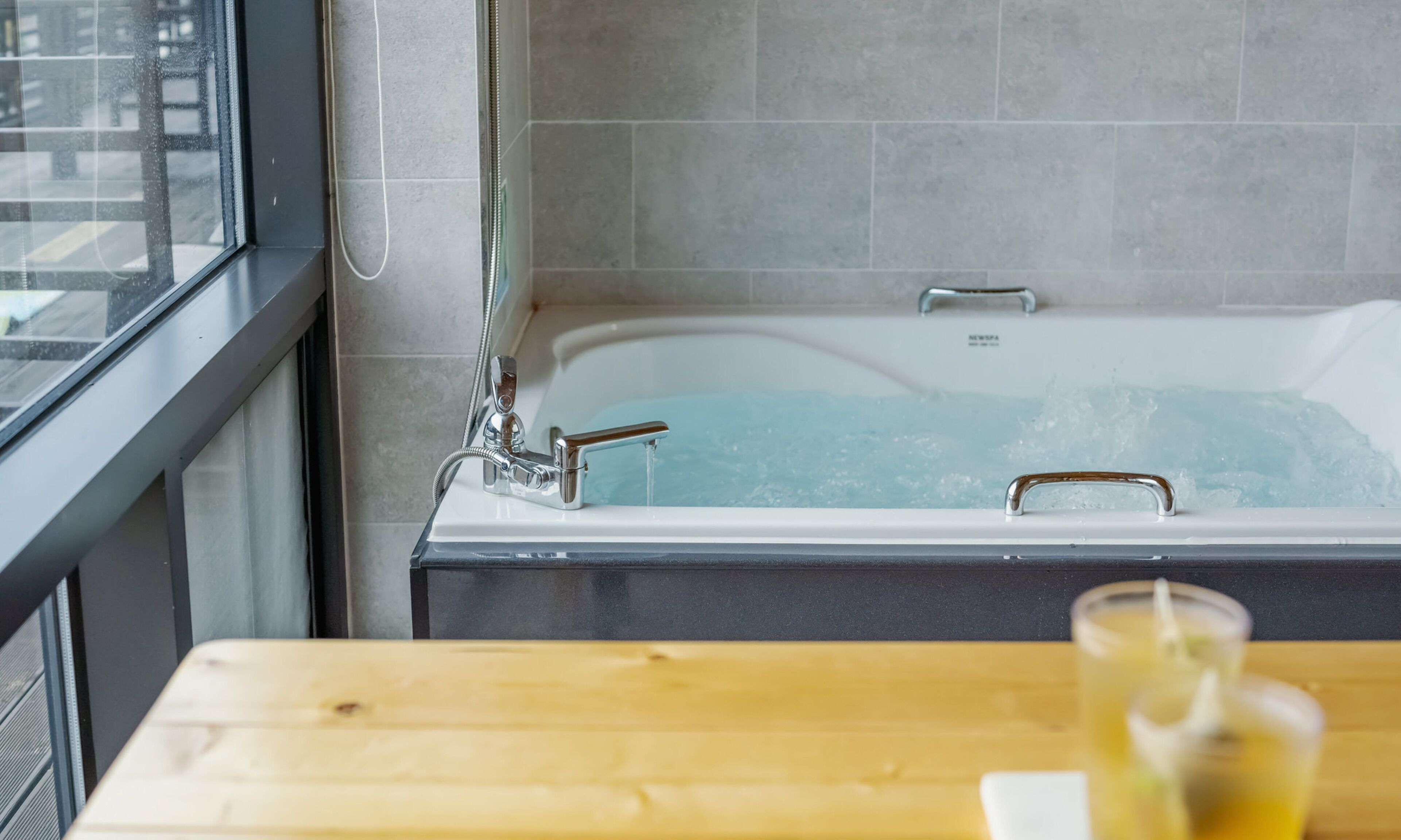 Une salle de bain moderne avec une baignoire, une fenêtre et une table en bois sur laquelle se trouve un verre d’eau.