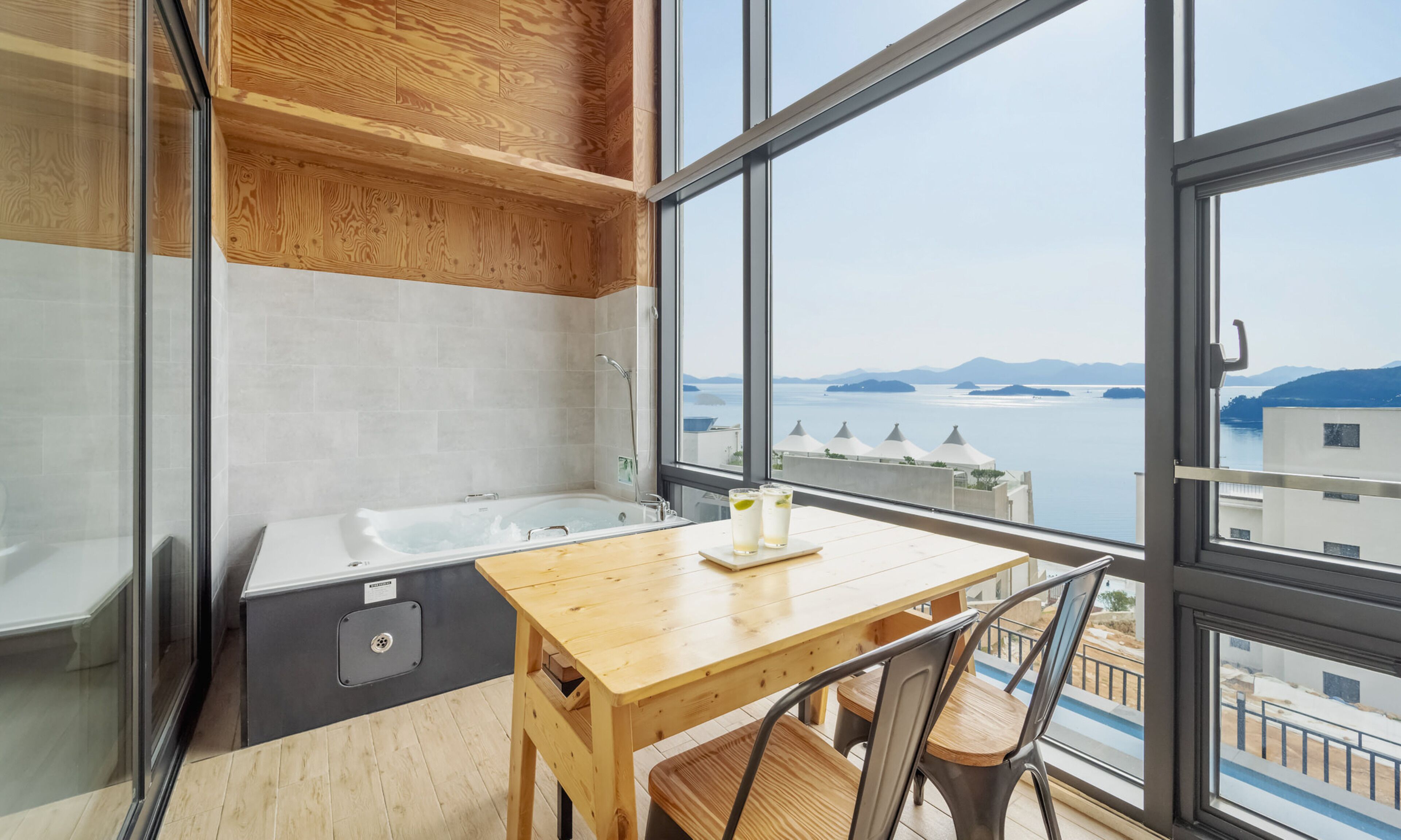 Une salle de bain moderne avec une baignoire, une table et des chaises en bois, ainsi qu’une grande fenêtre offrant une vue sur la mer et les bâtiments.
