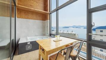 Une salle de bain moderne avec une baignoire, une table et des chaises en bois, ainsi qu’une grande fenêtre offrant une vue sur la mer et les bâtiments.