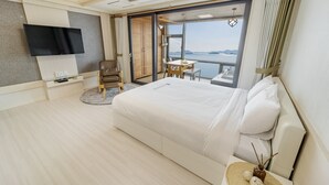 A modern hotel room with a large bed, a flat-screen TV, a small table with chairs, and a balcony with a view of the sea. - Yeosu Sunset Hostel and Pension (Yeosu)