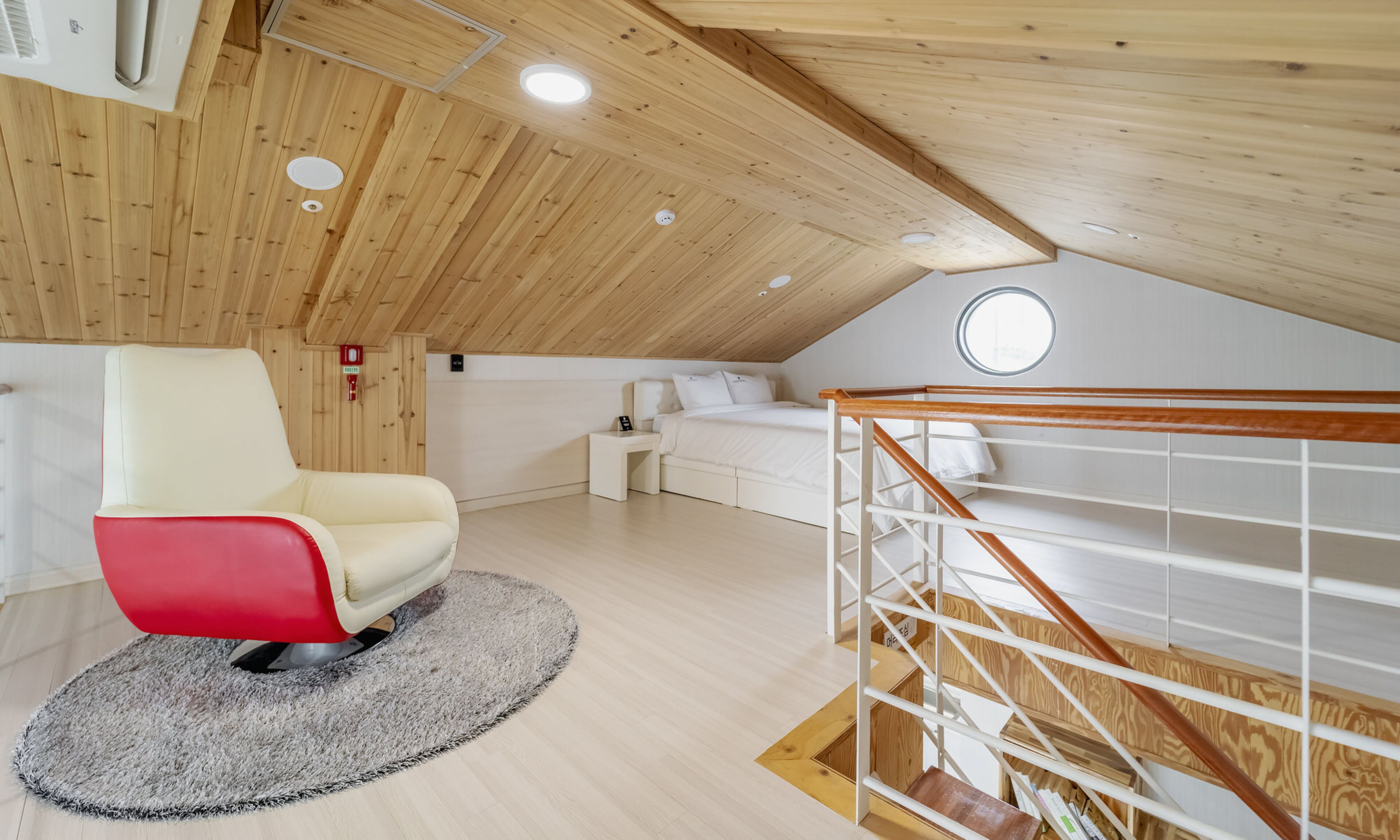 Ein modernes Schlafzimmer im Loft-Stil mit schrägem Dach, einem roten und weißen Stuhl, einem weißen Bett und einer Holztreppe.