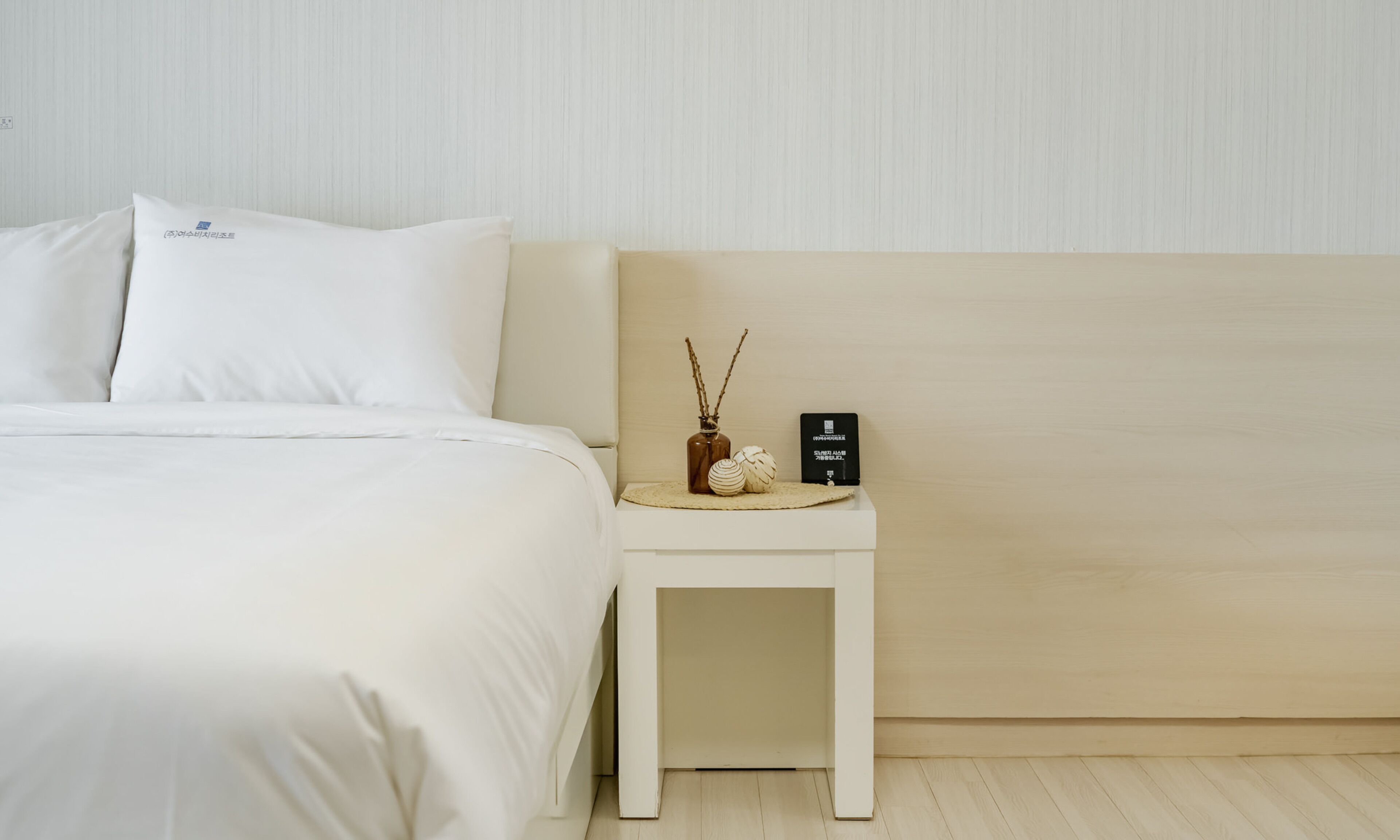 Un lit bien fait, avec du linge de lit blanc et des oreillers, une petite table de chevet agrémentée d’un objet décoratif et d’un objet noir, et une tête de lit de couleur claire.