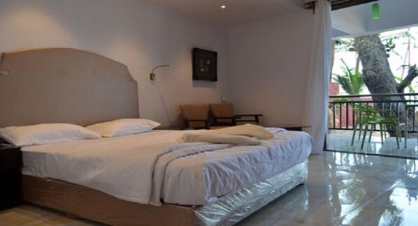 A hotel room with a large bed, a small table, a chair, and a balcony with a view of trees.
