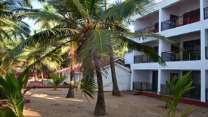 Un edificio de varios pisos con balcones, rodeado de palmeras y suelo arenoso.