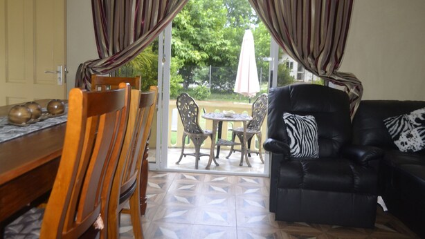 A living room with a black leather sofa, a wooden dining table with chairs, and a sliding glass door leading to an outdoor patio with a table and chairs. - My Ozi Perl Self Catering Guest House (Praslin Island)